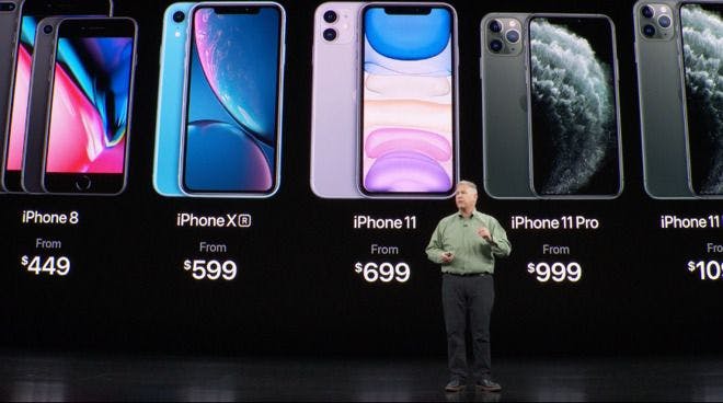 a man gives a public talk in front of a board displaying various models of iPhones, from the iPhone8 to the iPhone11, with their respective prices